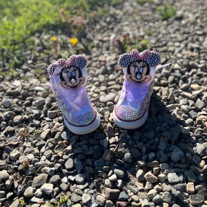 May include: A pair of custom-designed sneakers featuring Minnie Mouse. The shoes are white with a light purple translucent overlay, embellished with rhinestones and a Minnie Mouse face on the tongue. The shoes have a red trim and a white sole.