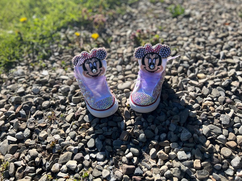 May include: A pair of custom-designed sneakers featuring Minnie Mouse. The shoes are white with a light purple translucent overlay, embellished with rhinestones and a Minnie Mouse face on the tongue. The shoes have a red trim and a white sole.