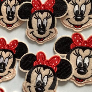 May include: Close-up of several Minnie Mouse patches. Each patch features Minnie's face with black ears, a red bow with white polka dots, and a smiling expression. The patches are beige with black and red accents.