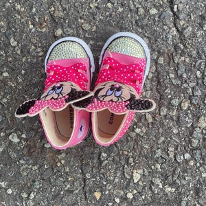 Puede incluir: Un par de zapatillas Converse rosas con suelas blancas y punteras con pedrer&iacute;a. Las zapatillas tienen cordones rosas de lunares y un dise&ntilde;o de Minnie Mouse en la leng&uuml;eta. Los zapatos son para ni&ntilde;os.