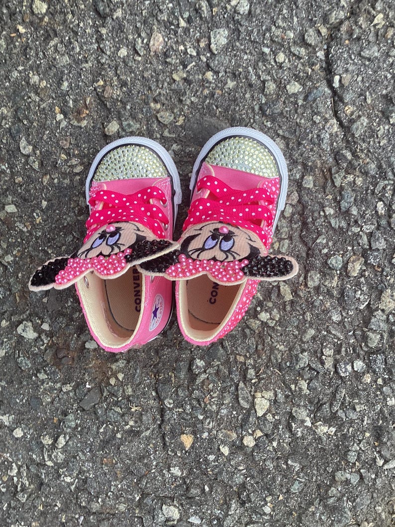 Puede incluir: Un par de zapatillas Converse rosas con suelas blancas y punteras con pedrer&iacute;a. Las zapatillas tienen cordones rosas de lunares y un dise&ntilde;o de Minnie Mouse en la leng&uuml;eta. Los zapatos son para ni&ntilde;os.