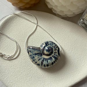 以下が含まれることがあります： 青と白の貝殻のデザインが施されたセラミックペンダントネックレス。ペンダントは繊細なシルバーチェーンから吊り下げられています。ネックレスは白いテクスチャのある表面に展示されており、背景にはキャンドルがあります。
