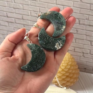 May include: Three crescent moon-shaped pendants made of green and white speckled stone. Each pendant has a silver-coloured bail for hanging. The pendants are held in a hand, with a brick wall in the background.
