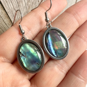 May include: Oval earrings featuring iridescent abalone shells set in silver-coloured metal frames. The earrings have a hook closure. The abalone shells display shades of green, blue, and purple. The earrings are displayed against a neutral background.