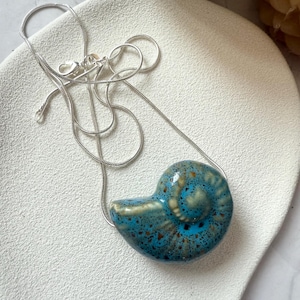 Op de afbeelding: Een ketting met een keramische hanger in de vorm van een blauw en bruin zeeschelp. De hanger hangt aan een fijne zilveren ketting. De zeeschelp heeft een glanzende afwerking. Op een witte achtergrond.
