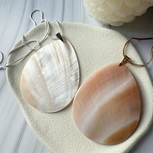 Puede incluir: Dos collares con colgantes en forma de lágrima. Uno es blanco nacarado, el otro melocotón y blanco. Ambos colgantes están sobre cadenas delicadas. Los collares descansan sobre una superficie texturizada blanquecina, sugiriendo una exhibición de joyas.