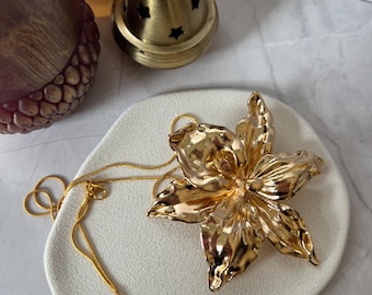 Puede incluir: Un collar de cadena dorada con un gran colgante de flor dorada. El colgante es una flor estilizada con una superficie texturizada.