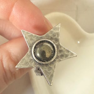 May include: A silver star ring with a hammered finish. The ring has a central circular setting containing a dark, iridescent stone. A distinctive piece of jewellery.
