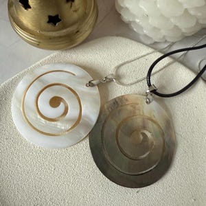 Könnte beinhalten: Zwei Muschelanhänger mit Spiraldesign, einer weiß und einer braun, an einer silbernen Kette und einer schwarzen Schnur.