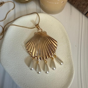 Puede incluir: Collar con colgante de concha dorada con cuentas colgantes de lágrima nacaradas. El colgante tiene un diseño texturizado en forma de abanico. El collar se muestra sobre una superficie blanca texturizada.