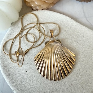 May include: A gold-colored necklace featuring a large, detailed seashell pendant. The pendant has a textured, fan-like design. The chain is a delicate, thin gold color. The necklace is displayed on a light-colored, textured surface.
