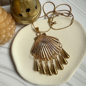 May include: A gold-colored necklace with a seashell pendant and teardrop dangles. The pendant is fan-shaped with vertical lines, and the teardrops hang below. The necklace is displayed on a cream-colored plate.