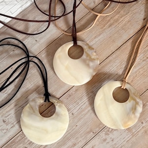 May include: Three circular pendants made of polished, cream-coloured stone with natural marbling, each featuring a central circular cutout. Each pendant is suspended from a long, thin cord in black, dark brown, or light tan, creating a set of necklaces.