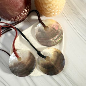 Könnte beinhalten: Drei schillernde Muschelanhänger mit braunen, schwarzen und roten Kordeln. Jeder Anhänger hat eine kreisförmige Form mit einem Loch in der Mitte. Die Muscheln haben einen perlmuttartigen Glanz mit Braun-, Weiß- und Regenbogenfarben.