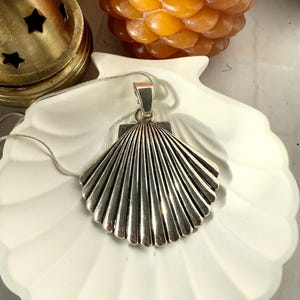 May include: A silver seashell pendant necklace on a white ceramic dish. The pendant is detailed with a textured, ribbed design.