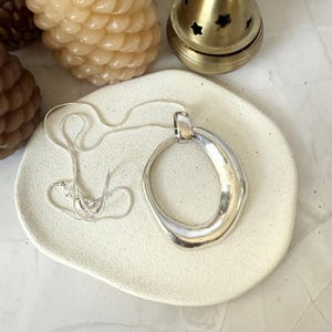 Könnte beinhalten: Eine silberne Halskette mit einem großen, abstrakten, ovalen Anhänger. Die Halskette wird auf einer weißen Keramikplatte präsentiert.