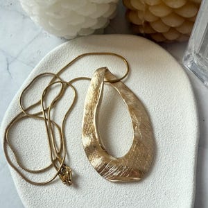 Puede incluir: Un collar dorado con un colgante texturizado en forma de lágrima. El colgante tiene un acabado de metal cepillado y cuelga de una cadena fina. El collar se exhibe sobre una superficie blanca, con velas en el fondo.