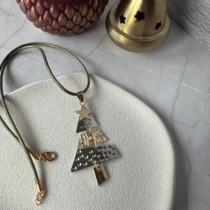 Könnte beinhalten: Eine Halskette mit einem Anhänger in Form eines Weihnachtsbaums mit einem goldenen Stern und geometrischen Formen in Gold und Silber. Die Halskette hat eine grüne Kordel mit einem goldenen Verschluss. Ein goldfarbener Kerzenhalter und eine kastanienbraune eichelförmige Kerze befinden sich im Hintergrund.