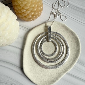 Puede incluir: Un collar plateado con tres colgantes circulares anidados. El collar descansa sobre una bandeja crema de forma orgánica. Dos velas decorativas están en el fondo. Una pieza de joyería.
