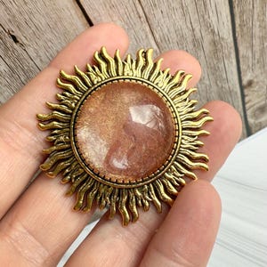 May include: A sun-shaped pendant with a bronze-coloured frame and a central, circular, pinkish-brown stone. The frame features stylised sun rays. The pendant is held in a hand, showcasing its size and design. The overall aesthetic is vintage and celestial.