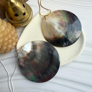 Puede incluir: Dos colgantes de concha iridiscente con cadenas plateadas y doradas. Las conchas muestran una mezcla de gris oscuro, negro y colores iridiscentes. También se ven un quemador de incienso dorado y una vela decorativa.
