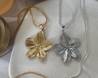 Puede incluir: Dos delicados collares de oro y plata con colgantes de flores. Los collares están sobre una superficie blanca.