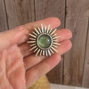 May include: A silver sunburst brooch featuring a central green gemstone. The brooch has radiating spikes around the gemstone, creating a sun-like design. The brooch is held in a hand against a wooden background.