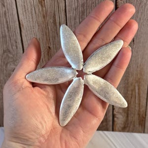 May include: A silver-coloured, flower-shaped decorative object with six elongated petals. The petals have a textured surface and are arranged around a central point. The object is held in a hand against a wooden background.
