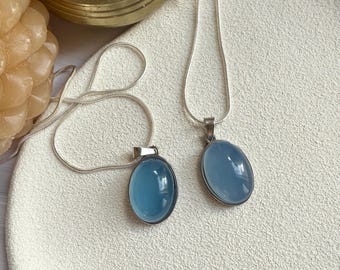 May include: Two silver necklaces with oval, light blue pendants. The pendants are set in silver bezels and hang from delicate silver chains. The necklaces rest on a white, textured surface, with a gold-colored metal container and a candle in the background.