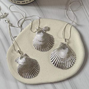 May include: Three silver seashell pendant necklaces displayed on a cream surface. Each necklace has a detailed, textured seashell pendant suspended from a delicate silver chain. The pendants are arranged to showcase their design, reflecting light.