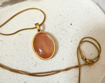 May include: A gold-toned necklace featuring an oval pendant with a peach-colored stone. The pendant is framed in gold and hangs from a delicate chain. The necklace is displayed on a white surface, showcasing its elegant design.