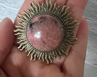 May include: A pink and black gemstone set in a gold-toned sunburst pendant.