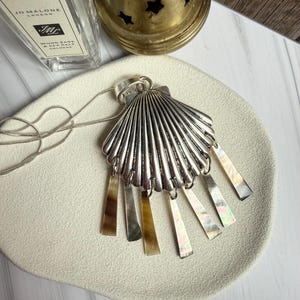 Puede incluir: Un collar con colgante de concha plateada con acentos rectangulares iridiscentes colgantes. El colgante está en una cadena plateada y descansa sobre un plato blanco texturizado. Una botella y un candelabro de latón están en el fondo.