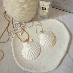 May include: Two seashell pendant necklaces on a white, textured, oval tray. One has a gold chain, the other a silver chain. A white candle and a Jo Malone London perfume bottle are in the background.