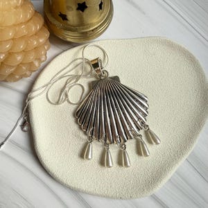 Puede incluir: Un collar con colgante de concha plateada con perlas de imitación colgantes. El colgante tiene un diseño detallado y acanalado y cuelga de una delicada cadena plateada. El collar se exhibe sobre una superficie de color crema.