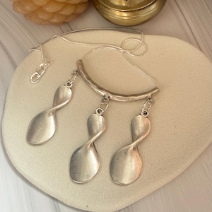 May include: A silver necklace with three abstract spoon-shaped pendants. The pendants hang from a curved bar and fine chain. The necklace is presented on a light-coloured, heart-shaped surface.