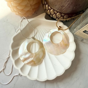 May include: Two iridescent shell pendant necklaces with silver and gold chains rest on a white seashell-shaped dish. The pendants have a circular design with a cutout center, showcasing a pearlescent sheen. A candle and a decorative item are in the background.