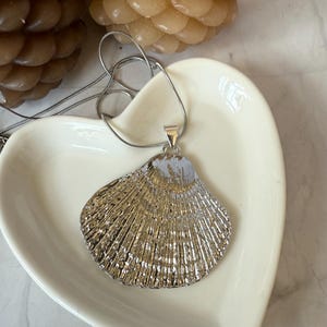Könnte beinhalten: Halskette mit silbernem Muschel-Anhänger. Der Anhänger hat ein strukturiertes, fächerförmiges Design mit einer silbernen Kette. Die Halskette liegt in einer weißen, herzförmigen Schale, mit zwei beigen Kerzen im Hintergrund.