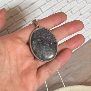 May include: An oval pendant featuring a polished stone with a mottled texture in shades of deep red, dark green, and grey. The stone is set within a smooth, metallic silver-toned frame and includes a small silver-toned bail at the top for a chain.