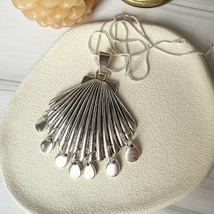 Op de afbeelding: Een zilverkleurige hangerketting met een waaierontwerp en bungelende cirkelvormige bedels. De hanger is bevestigd aan een fijne zilveren ketting. De ketting wordt gepresenteerd op een crèmekleurig oppervlak.