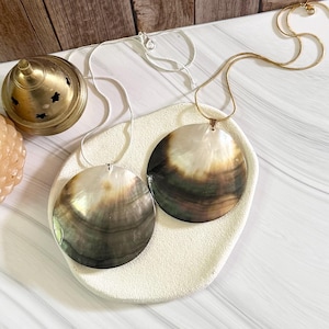 Könnte beinhalten: Zwei runde, schillernde Muschel-Anhänger-Halsketten. Eine Halskette hat eine silberne Kette, die andere eine goldene Kette. Die Anhänger sind dunkel mit einem perlmuttartigen Glanz. Die Halsketten liegen auf einer weißen, strukturierten Oberfläche.