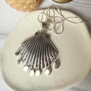 Puede incluir: Un collar con colgante plateado con un diseño en forma de abanico y colgantes circulares colgantes. El colgante está unido a una delicada cadena plateada. El collar se exhibe sobre una superficie color crema.