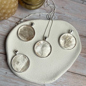 May include: Four circular pendants with a shimmering, textured off-white inlay set in polished silver-toned metal frames. One pendant is attached to a thin silver-toned chain, while the other three are separate, all displayed on a textured surface.