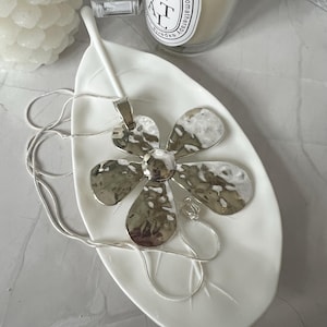 Könnte beinhalten: Eine silberne Blumenkette auf einer weißen, blattartigen Schale. Der Anhänger ist eine gehämmerte Metallblume mit fünf Blütenblättern.