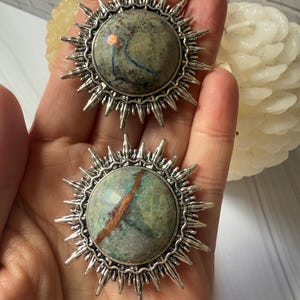 Puede incluir: Dos colgantes en forma de sol con un marco plateado y una piedra central. Las piedras tienen una mezcla de colores verdes, azules y marrones. Los colgantes se sostienen en una mano.