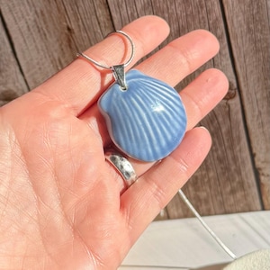 May include: A light blue seashell pendant with a silver chain. The pendant is a ceramic shell with detailed ridges. The necklace is held in a hand, with a silver ring on the finger.