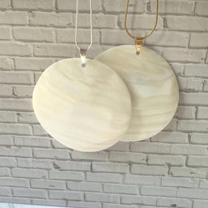 May include: Two round, cream-colored pendant necklaces. One has a silver chain, the other a gold chain. The pendants have a marbled appearance. The background is a light gray brick wall.