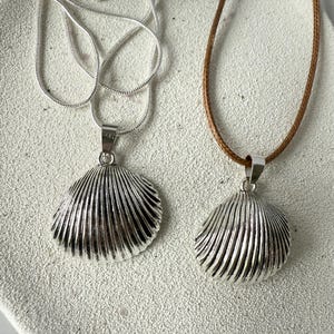 May include: Two seashell pendant necklaces. One has a silver chain, the other a brown cord. The pendants are silver-coloured, with a ribbed texture. The necklaces are displayed on a light-coloured surface.