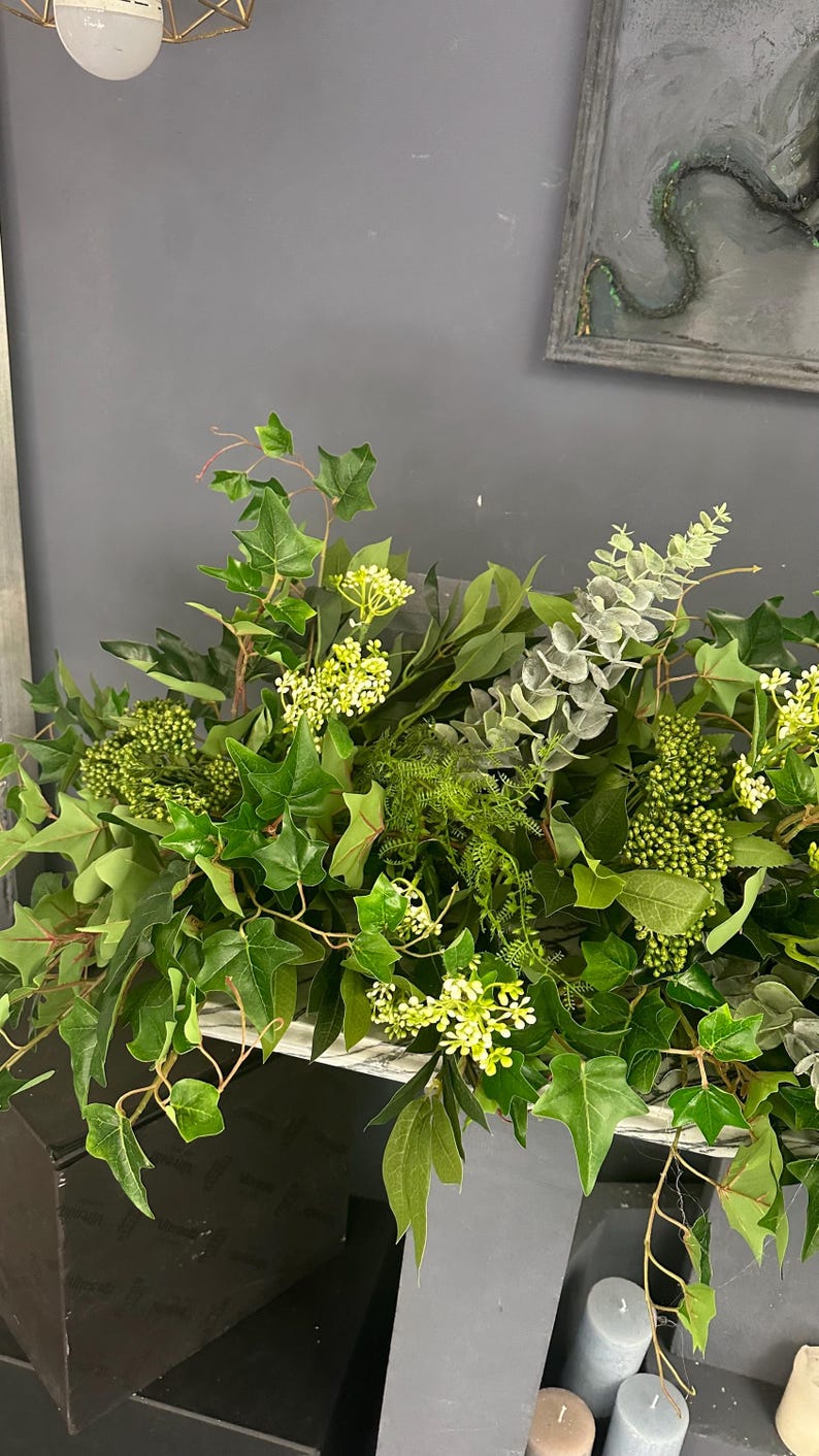 Pode incluir: Uma guirlanda verde exuberante com uma mistura de folhas, videiras e pequenas flores brancas. A guirlanda est&aacute; drapeada sobre uma superf&iacute;cie cinza e &eacute; acentuada por velas cinzas.