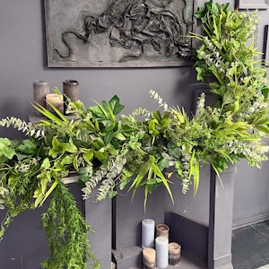 May include: A gray fireplace mantel decorated with a green garland of eucalyptus, ivy, and other greenery. The mantel has a gray background and is decorated with candles.